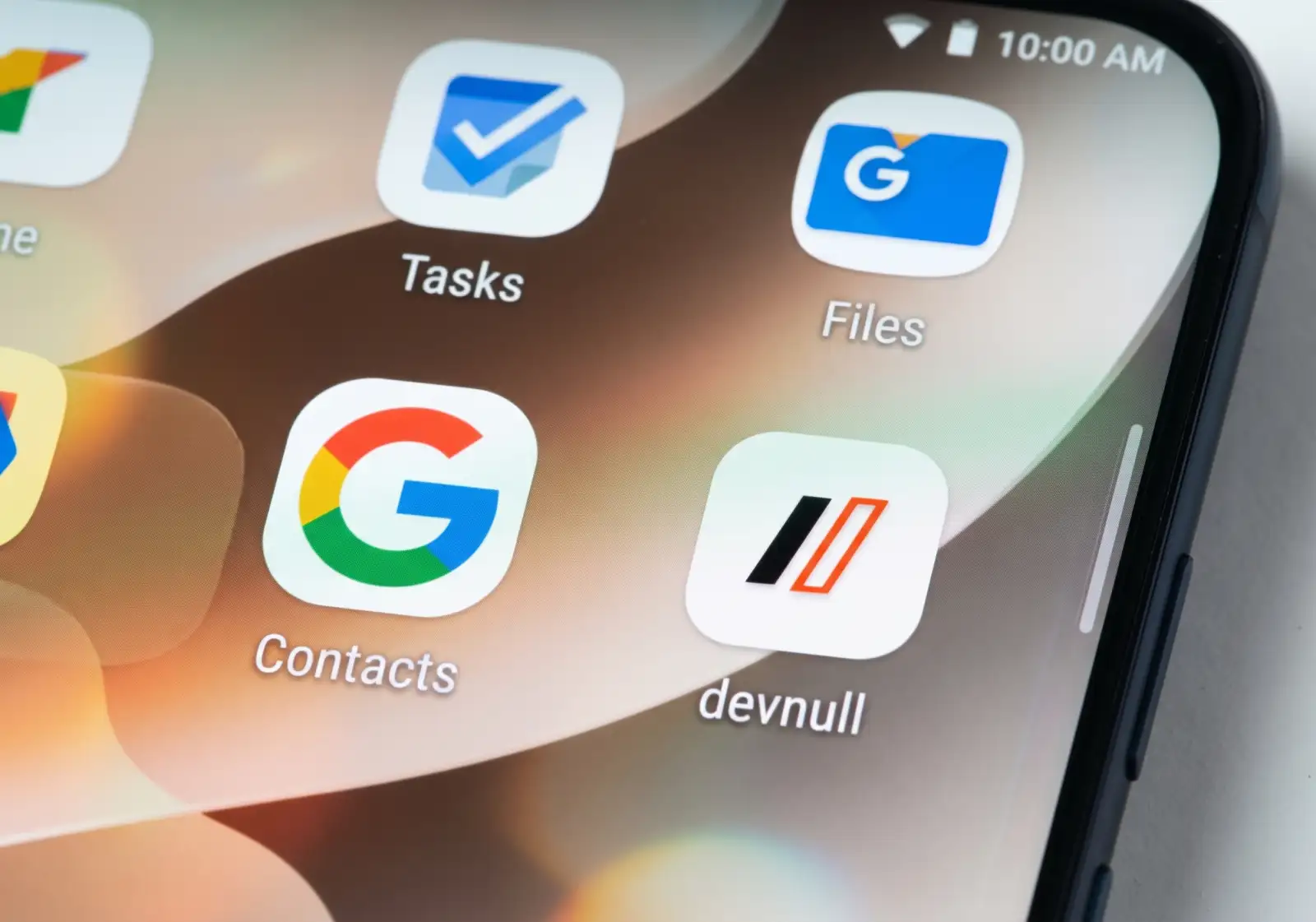 A macro photograph of a modern Android smartphone screen, showing a grid of app icons on a blurred light-brown gradient wallpaper. Prominently displayed are the Google Tasks, Google Files, and Google Contacts icons. The custom 'devnull' icon from the original image is preserved in the lower right, featuring a black and orange geometric logo on a white squircle. Standard Android status bar icons are visible at the top. The phone's curved edge is visible on the right.